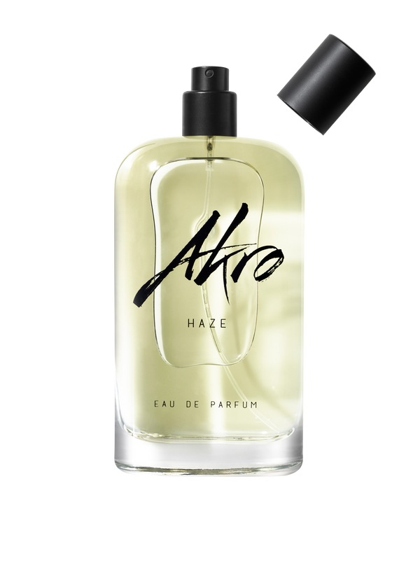 A clear glass bottle of Akro HAZE Eau de Parfum, adorned with black text on the front, showcases a light yellow liquid inside. Topped with a black cap, this addictive chill fragrance exudes a fresh smoky scent that intrigues and captivates.