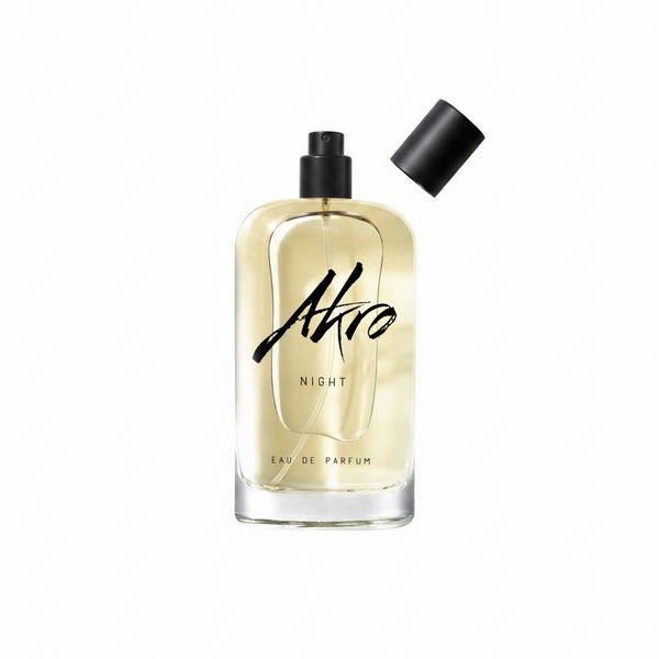 A bottle of NIGHT by Akro. The transparent container, accompanied by a black cap placed aside, reveals a spray nozzle, while the light amber liquid inside suggests musky notes and Bulgarian rose essences.
