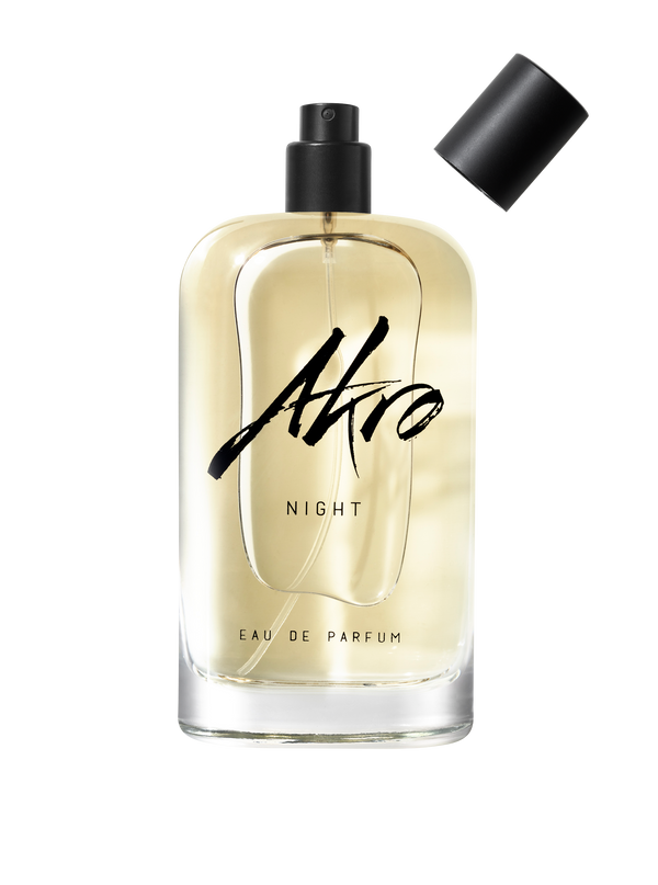 A bottle of NIGHT by Akro. The transparent container, accompanied by a black cap placed aside, reveals a spray nozzle, while the light amber liquid inside suggests musky notes and Bulgarian rose essences.