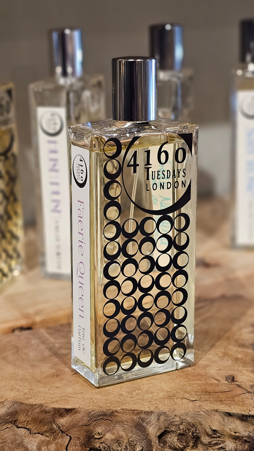 A perfume bottle labeled "4160Tuesdays" with "Faerie Queen" on the side, displayed on a wooden surface with other blurred perfume bottles in the background. The elegant scent whispers of vanilla and kumquat, adding a touch of magic to your daily routine.
