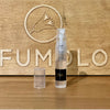 A 2ml transparent spray vial with a black label sits on a wooden surface. The wooden background has an engraved bird and the partial word "PERFUMOLO." The vial's cap is removed and placed beside it, hinting at the aromatic blend of Erba Pura by Xerjoff captured inside.