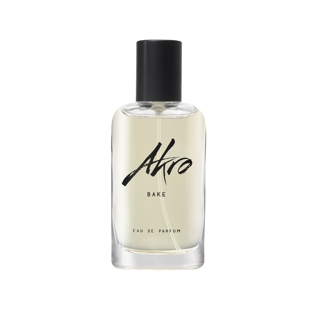 A clear bottle of BAKE Eau de Parfum by Akro features a black cap and a minimalist design. The label showcases the brand name "Akro" and the product name "BAKE" in black text, hinting at a delightful lemon cupcake fragrance.