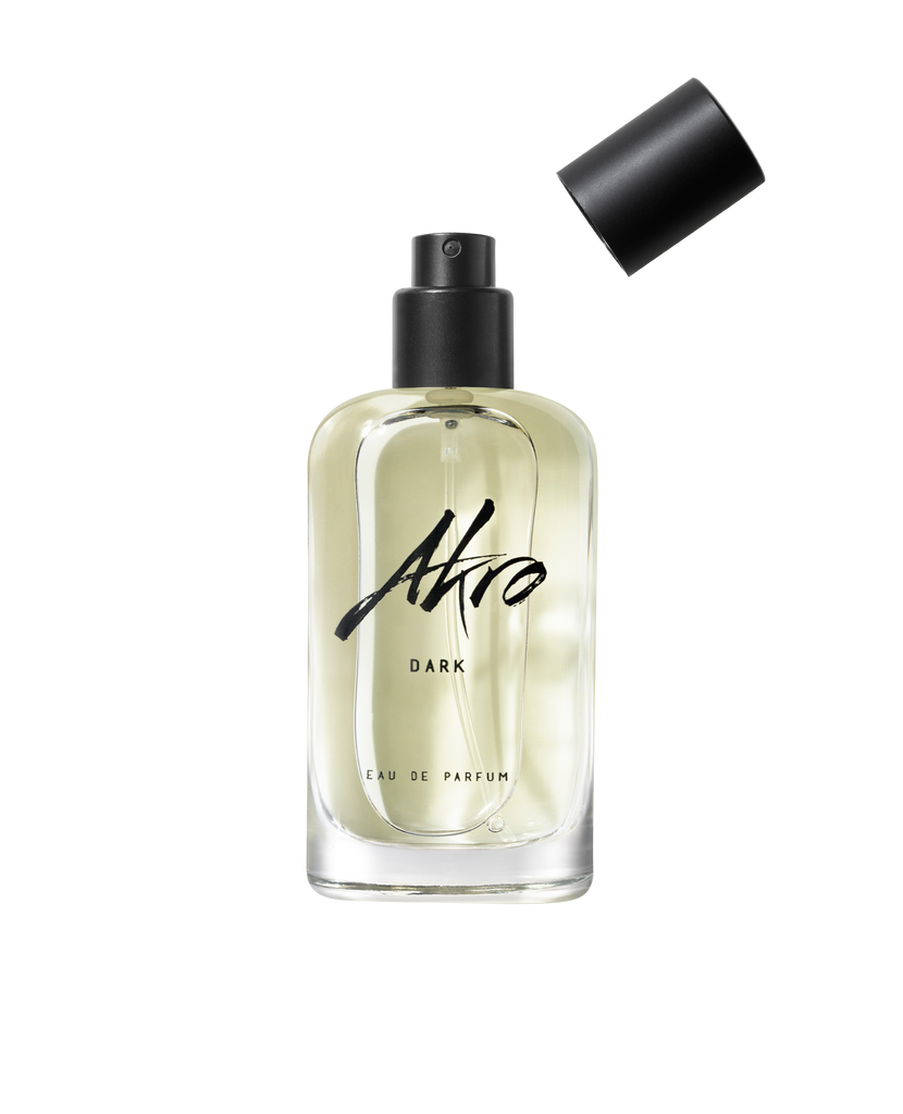 A clear glass bottle of Akro DARK Eau de Parfum featuring a black spray nozzle, with the cap placed nearby. The bottle is partially filled with a light-colored liquid, capturing the allure of mature sensuality intertwined with an addictive dark chocolate fragrance.