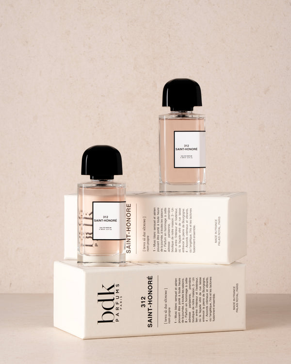 Two 312 Saint-Honoré by BDK Parfums bottles with black caps are elegantly positioned atop their respective white packaging boxes on a beige surface, exuding notes of orange blossom and white musks.
