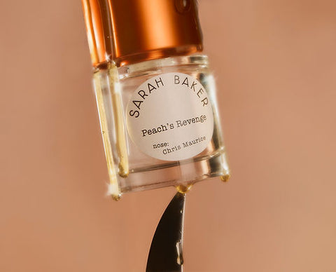 A close-up image of a perfume bottle labeled "Sarah Baker Peaches Revenge." The bottle is upside down, and some liquid is dripping from it. The label also mentions "nose: Chris Maurice." A hint of peach and spices subtly lingers around the scene.