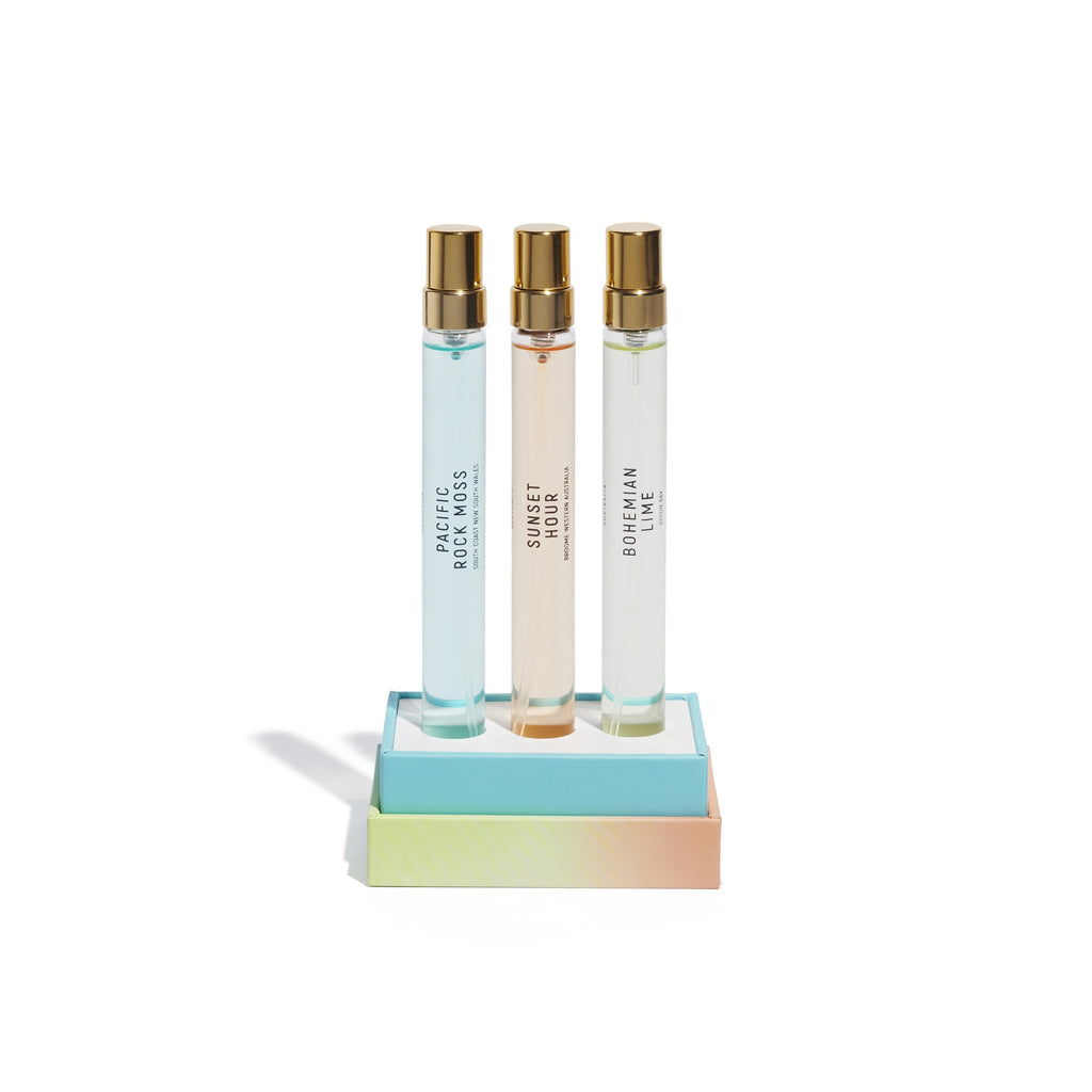 Three perfume bottles, featuring "Pacific Rock Moss," "Sunset Hour," and "Bohemian Lime," are elegantly arranged on a gradient-colored stand, epitomizing the luxurious Coast to Coast collection from Goldfield & Banks.