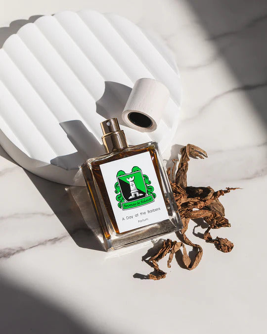 A perfume bottle labeled "A Day at the Barbers" by Aromas de Salazar with a green cat logo lies on a white surface next to dried plant leaves and a textured white background element, exuding a luxurious scent reminiscent of a traditional barbershop experience.