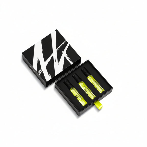 The AKRO 3 x 2ml Discovery Kit by Akro comes in a sleek black box featuring a distinctive white "W" design. Inside, three yellow spray bottles of exquisite fragrances are securely placed in foam cutouts, with one bottle lying next to the box.