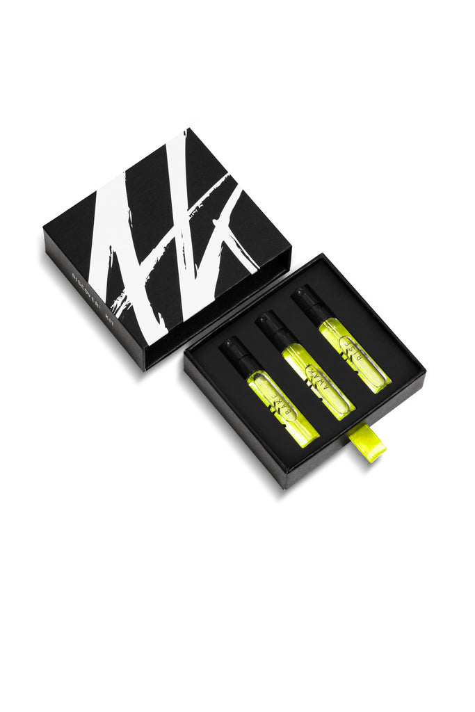 The AKRO 3 x 2ml Discovery Kit by Akro comes in a sleek black box featuring a distinctive white "W" design. Inside, three yellow spray bottles of exquisite fragrances are securely placed in foam cutouts, with one bottle lying next to the box.