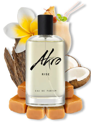 A bottle of RISE by Akro Eau de Parfum surrounded by caramel cubes, a coconut half, a vanilla flower, and a coconut drink with a straw and mint garnish—capturing the essence of summer fragrance with hints of sandalwood.