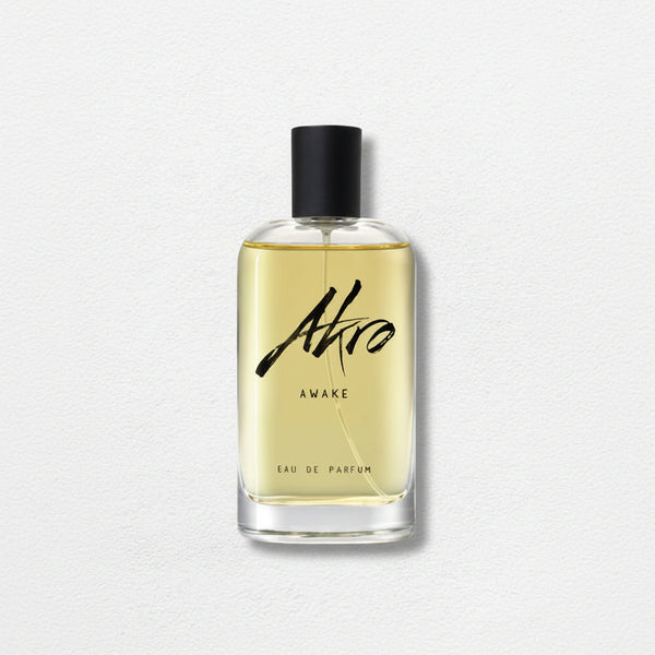 A bottle of Akro AWAKE Eau de Parfum with a black cap and clear glass container filled with light yellow liquid. The label features black cursive text, capturing the essence of a coffee fragrance in this unisex perfume.