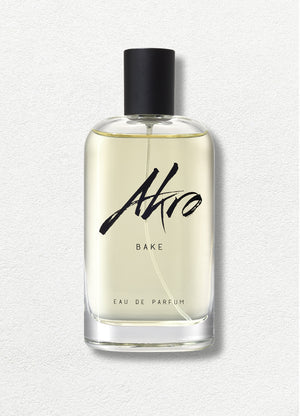 A clear bottle with a black cap contains a pale yellow liquid labeled "BAKE" by Akro in black cursive text on the front, evoking a delectable lemon cupcake fragrance.
