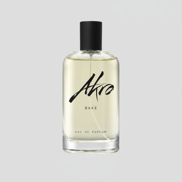 A clear bottle with a black cap contains a pale yellow liquid labeled "BAKE" by Akro in black cursive text on the front, evoking a delectable lemon cupcake fragrance.