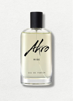 A clear glass bottle of "RISE" by Akro Eau de Parfum, featuring a black cap and bold, black script text on a light background, captures the essence of summer fragrance with hints of Coconut Water and Sandalwood.