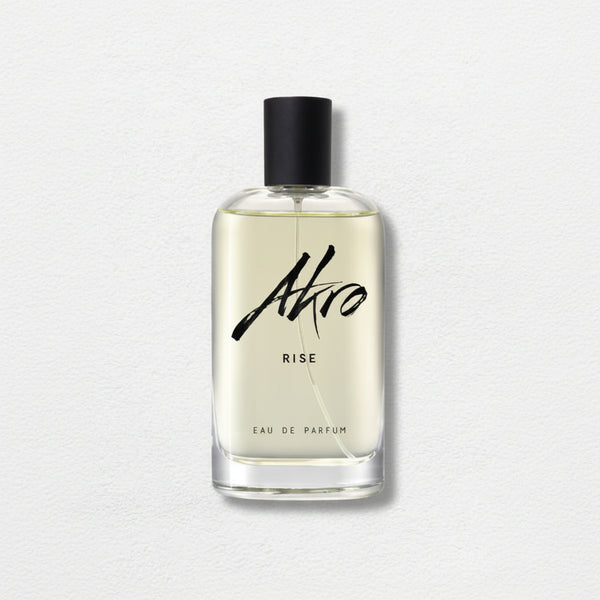 A clear glass bottle of "RISE" by Akro Eau de Parfum, featuring a black cap and bold, black script text on a light background, captures the essence of summer fragrance with hints of Coconut Water and Sandalwood.