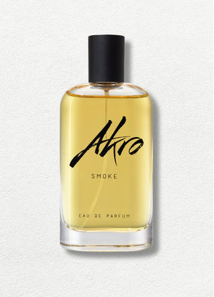A clear glass bottle of Akro's SMOKE Eau de Parfum, featuring a black cap and filled with amber-colored liquid, exudes a rich tobacco fragrance. The product is set against a light textured background.