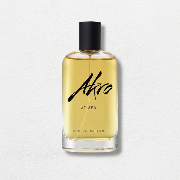 A clear glass bottle of Akro's SMOKE Eau de Parfum, featuring a black cap and filled with amber-colored liquid, exudes a rich tobacco fragrance. The product is set against a light textured background.