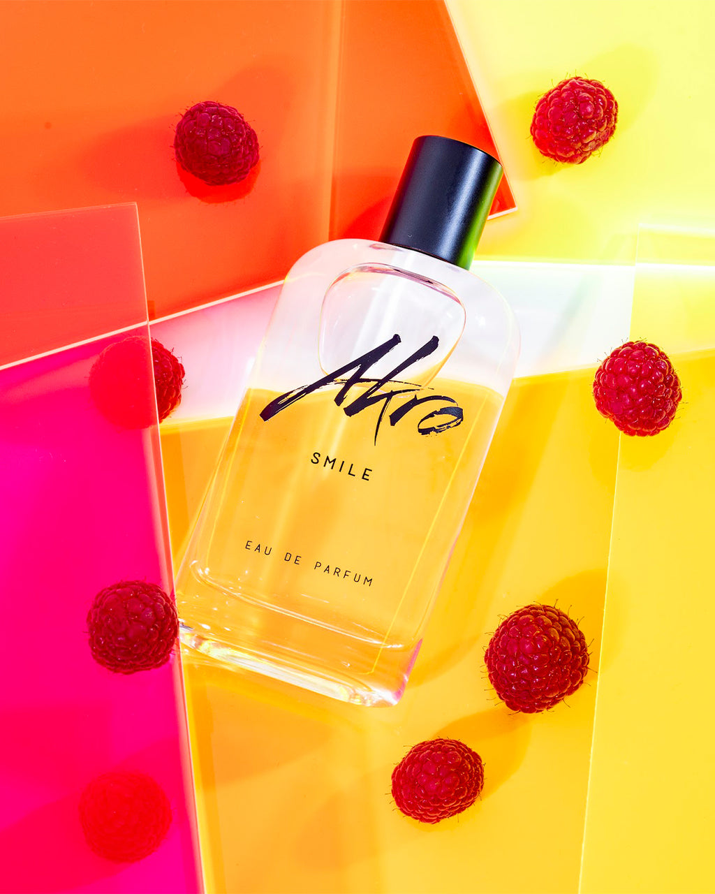 A bottle of "Akro SMILE" lies on a colorful background with red raspberries scattered around, embodying the essence of a unisex fragrance and encapsulating the joyous spirit of happiness.