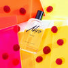 A bottle of "Akro SMILE" lies on a colorful background with red raspberries scattered around, embodying the essence of a unisex fragrance and encapsulating the joyous spirit of happiness.