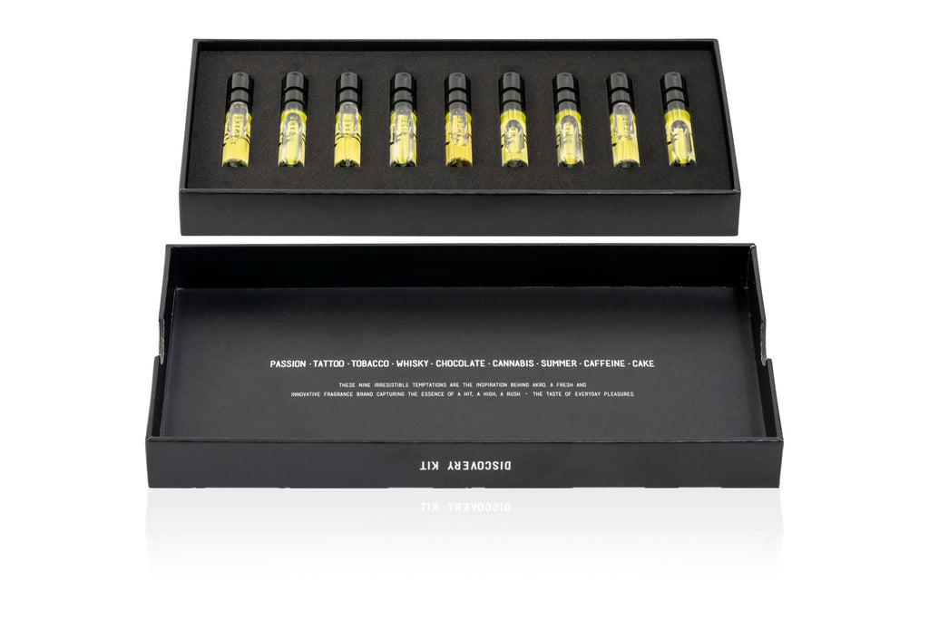 An open black box from the AKRO 9 x 2ml Discovery Kit by Akro reveals a collection of nine small vials filled with yellow liquid, neatly organized in a foam insert. The lid of the box features various words such as "passion," "chocolate," and "caffeine." These bold fragrances turn addictions into perfumes, promising an enticing olfactory experience.