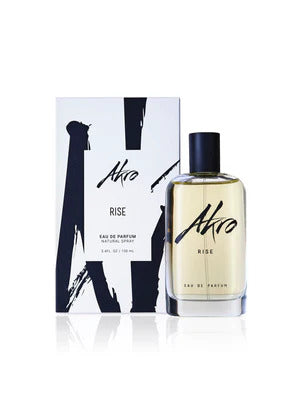 A bottle of Akro RISE Eau de Parfum, an ideal summer scent, is displayed in front of its coordinating packaging. The clear glass bottle with a black cap encases the perfume, while the packaging features a stylish white and black design.