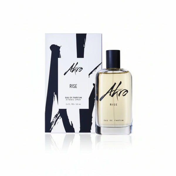 A bottle of Akro RISE Eau de Parfum, an ideal summer scent, is displayed in front of its coordinating packaging. The clear glass bottle with a black cap encases the perfume, while the packaging features a stylish white and black design.