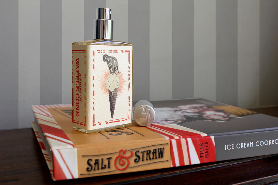 A bottle of Imaginary Authors' A Whiff of Wafflecone rests on top of two books, "Ice Cream Cookbook" by Tyler Malek and another with the same branding from an ice cream shoppe. An LA cap pin lies nearby, leaving you wondering, Howdtheydoit?