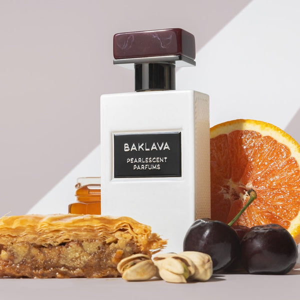 A bottle of Baklava by Pearlescent Parfums is placed beside a slice of baklava, some nuts, cherries, honey, and a half sweet orange, all set against a minimalist background.