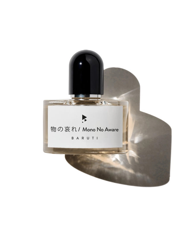 A clear glass bottle of "Mono No Aware" perfume by Baruti, adorned with a black cap and a white label showcasing Japanese characters and English text. Enhanced by cherry blossom notes, the bottle stands against a white background with its shadow subtly visible.