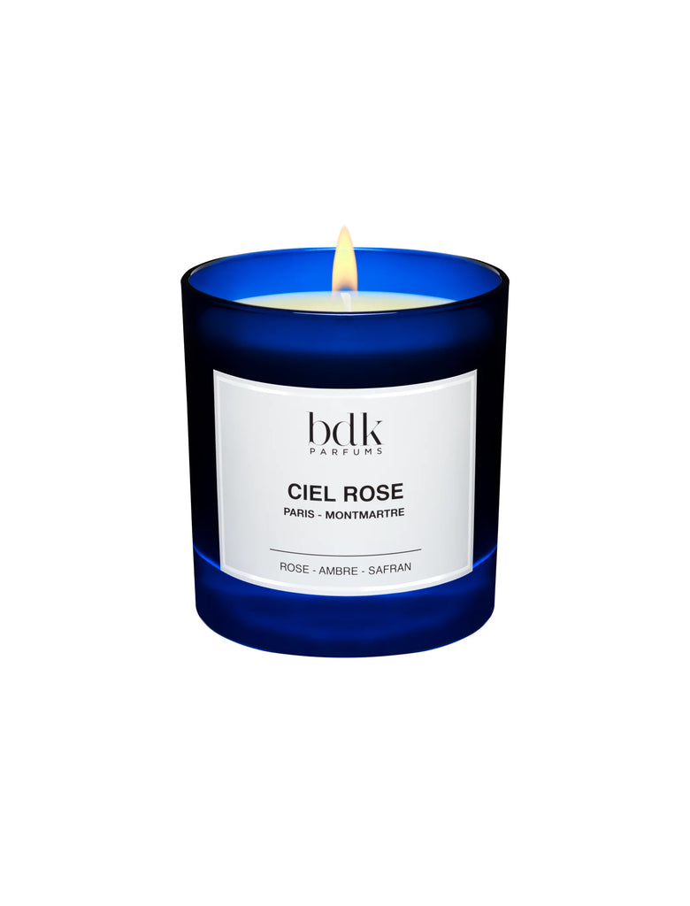 A lit candle in a dark blue glass container labeled "BDK Parfums Ciel Rose." The label indicates "Rose - Ambre - Safran," while the ambiance is enhanced with the delicate scent of fresh rose and a hint of white musk.