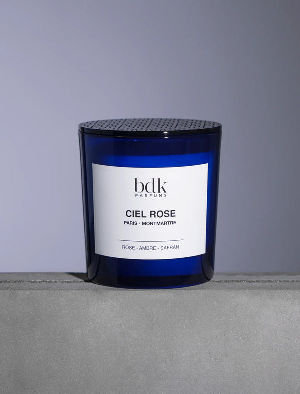 A blue glass candle labeled "Ciel Rose" by BDK Parfums, featuring notes of fresh rose, amber, and saffron accord, sits on a gray surface.