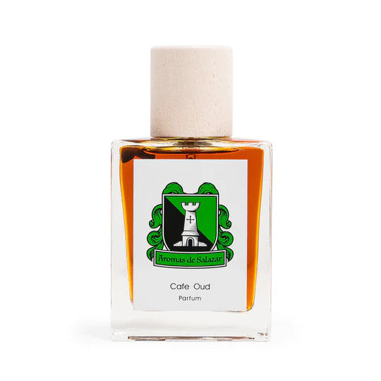 A clear glass bottle of "Cafe Oud" perfume from Aromas de Salazar, filled with amber-colored liquid and capped with a beige top. The label displays a green emblem featuring a tower and a cross, showcasing its luxurious blend of Cambodian oud and dark aged patchouli.