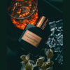 Positioned next to a glass of dark, ice-filled liquid, some loose ice cubes, and delicate white flowers lies a bottle of Chris Collins' "Harlem Nights" eau de parfum—an evening fragrance that whispers seduction.
