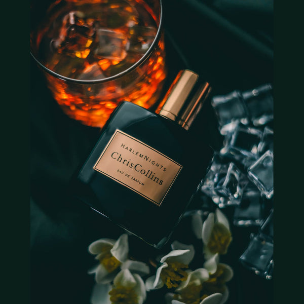 Positioned next to a glass of dark, ice-filled liquid, some loose ice cubes, and delicate white flowers lies a bottle of Chris Collins' "Harlem Nights" eau de parfum—an evening fragrance that whispers seduction.