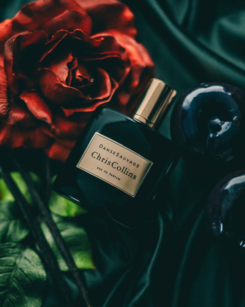 A bottle of Danse Sauvage by Chris Collins sits on a black satin surface, exuding a sensual scent, accompanied by a red rose and dark decorative elements.