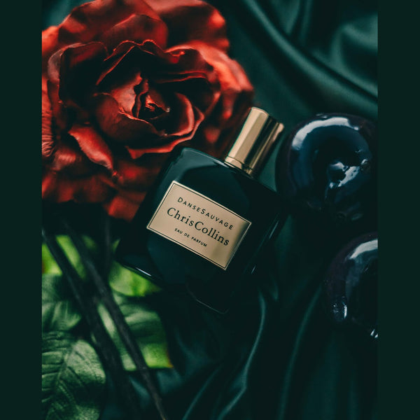 A bottle of Danse Sauvage by Chris Collins sits on a black satin surface, exuding a sensual scent, accompanied by a red rose and dark decorative elements.
