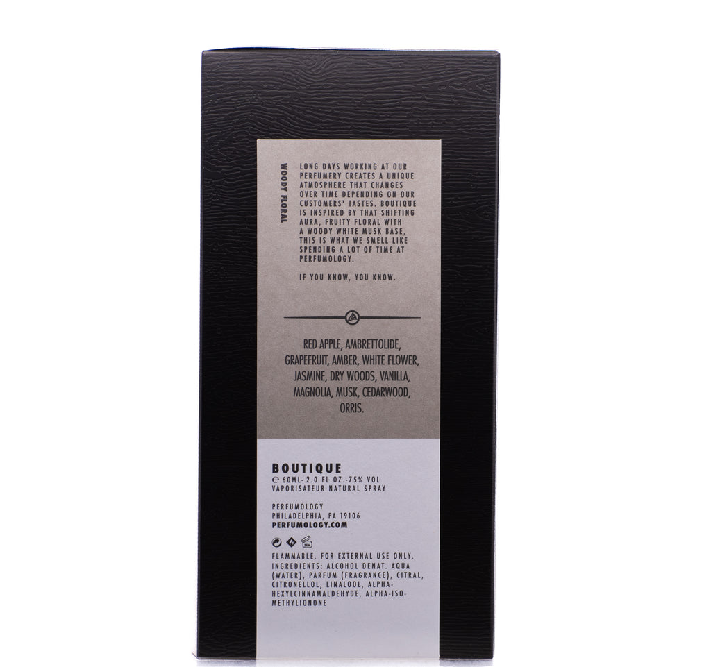 A brown box with a detailed label listing ingredients such as red apple, ambrette, and cedarwood. The fruity floral description highlights its blend of scents, including woody white musk. Usage instructions include warnings and perfume information from Perfumology's Boutique in Philadelphia, PA.