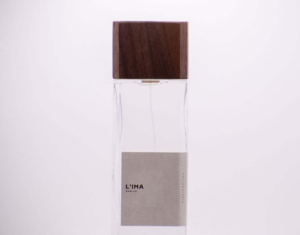 A clear rectangular perfume bottle from Perfumology with a wooden cap and a beige label that reads "L'Ima," exuding hints of citrus notes and floral undertones. The background is plain white.