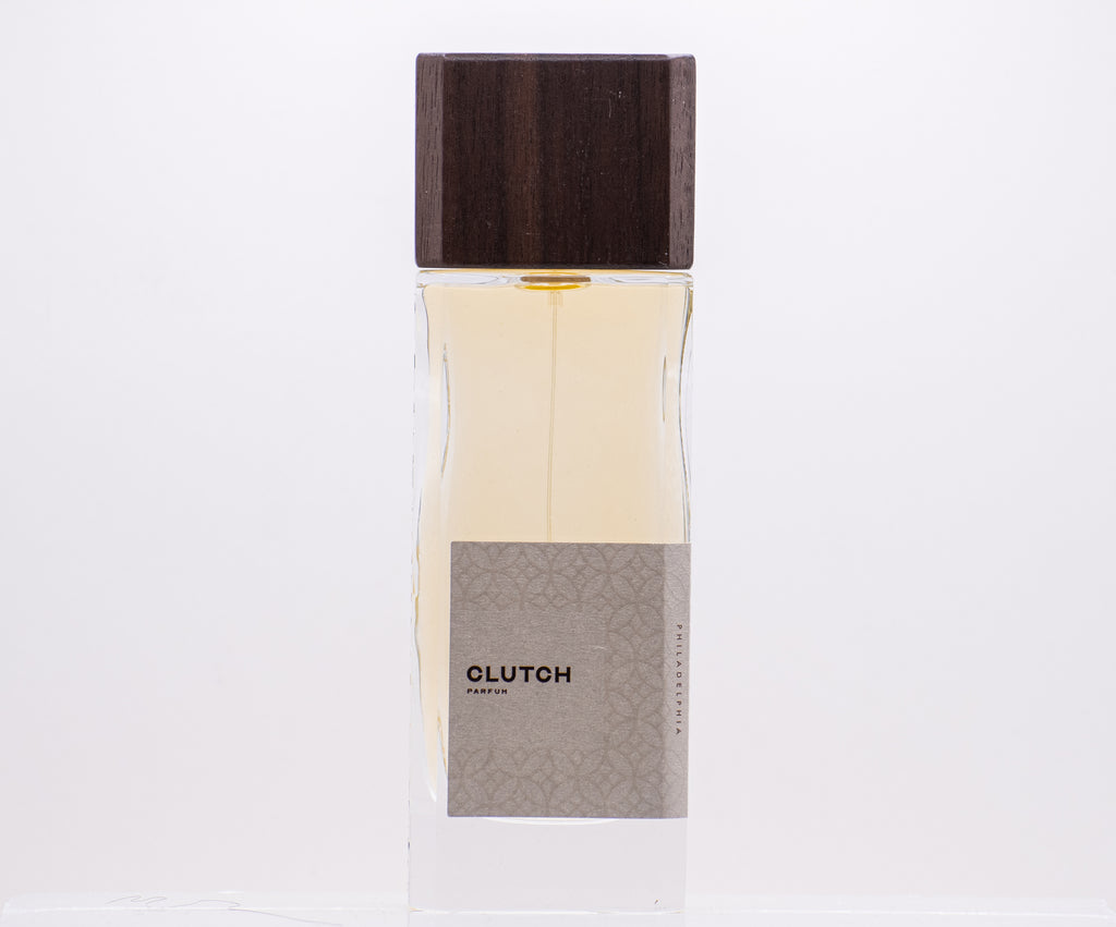 Clear glass bottle with yellow liquid, a brown wooden cap, and a label reading "CLUTCH PARFUM" in black text on the front. This independent perfume from Perfumology boasts an exhilarating scent that is sure to captivate.