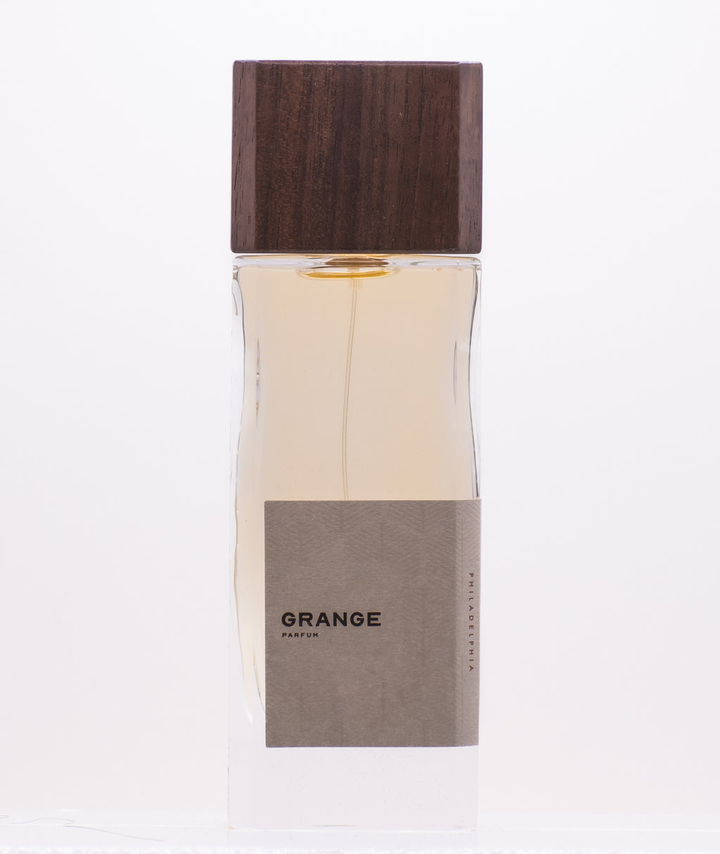 A bottle of perfume labeled "Grange by Perfumology" with a wooden cap and a light brown liquid inside, composed by Justin Frederico to reflect the essence of this fresh creek experience and evoke nostalgia for moments in the woods during childhood.