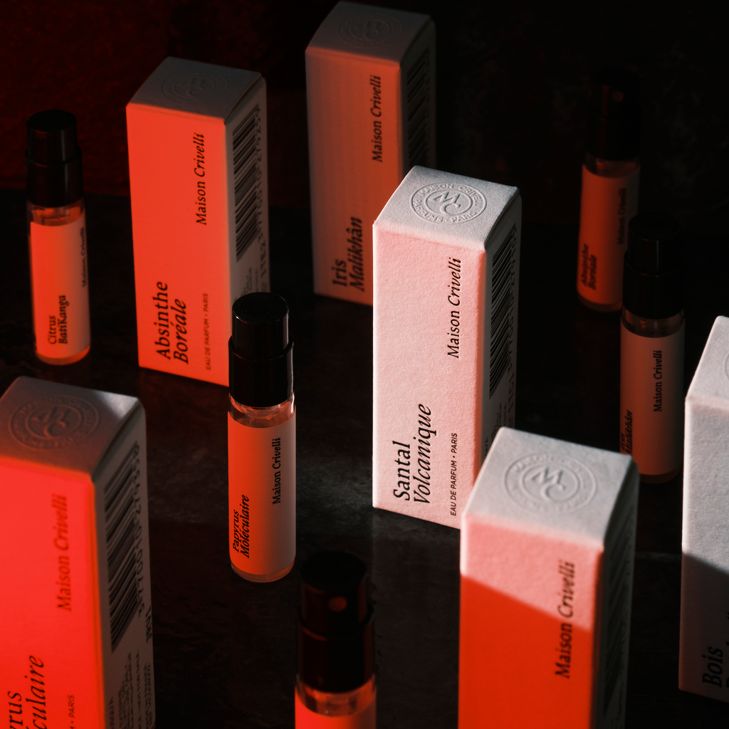 Several perfume vials and boxes with various labels are arranged on a surface under dramatic red and black lighting, showcasing the Maison Crivelli Discovery Set, 10x1.5ml, and its unique fragrances.