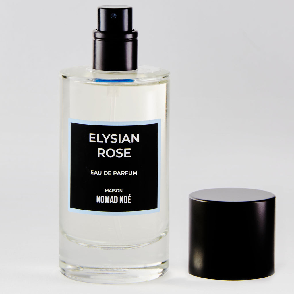 A clear glass bottle of Elysian Rose eau de parfum by Nomad Noe, showcasing a modern rose scent with a black label, black spray nozzle, and a black cap placed beside it.
