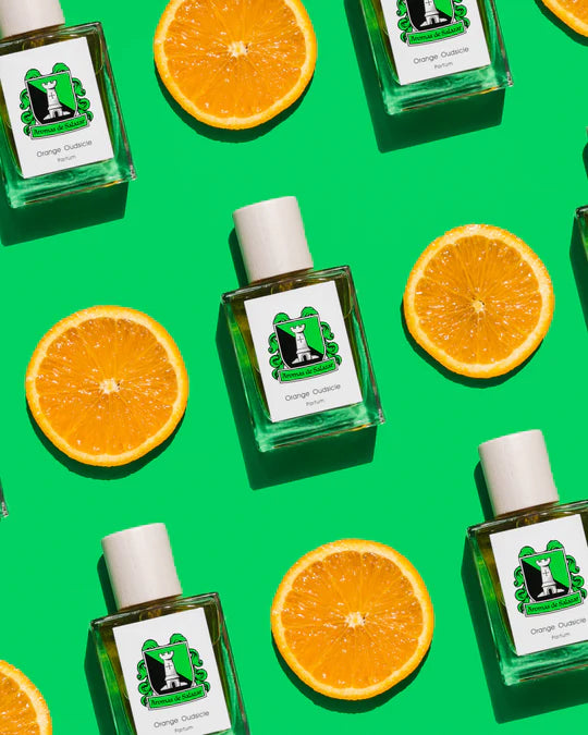 Bottles of Aromas de Salazar's "Orange Oudsicle" and halved orange slices are arranged in a diagonal pattern on a bright green background.