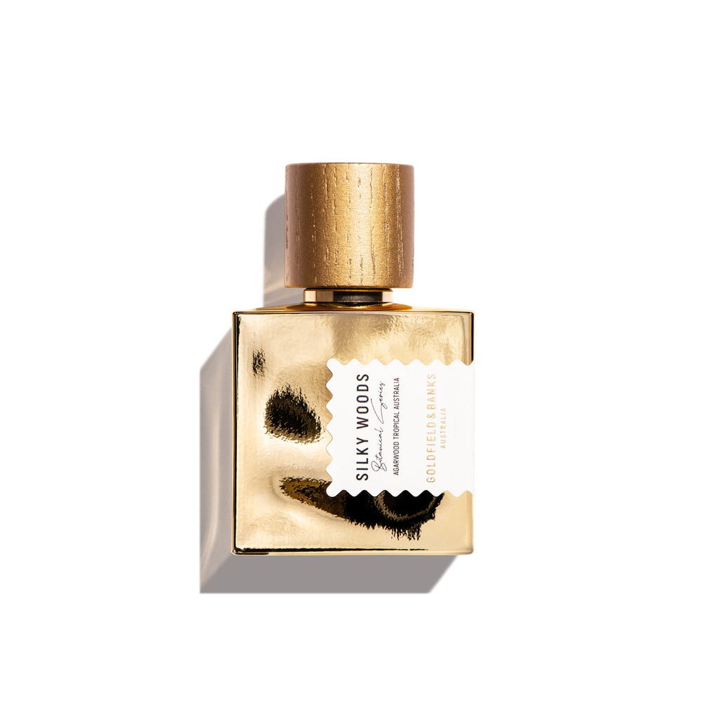 A rectangular perfume bottle labeled "Silky Woods" by Goldfield & Banks, designed with a gold finish and a textured cap, featuring an irregularly placed label, stands elegantly against a white background.