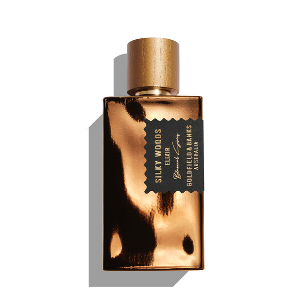 A metallic gold perfume bottle with a black label reading "Silky Woods Elixir by Goldfield & Banks." The bottle features a cylindrical gold cap, capturing the exotic botanical essence enriched with the luxuriousness of agarwood.