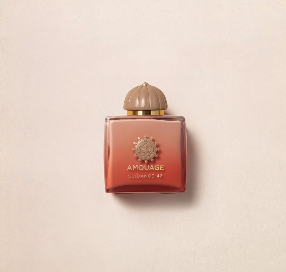 A square bottle of Amouage Guidance 46 perfume, featuring a gradient rose to brown color and topped with a round beige cap, is set against a light beige background.