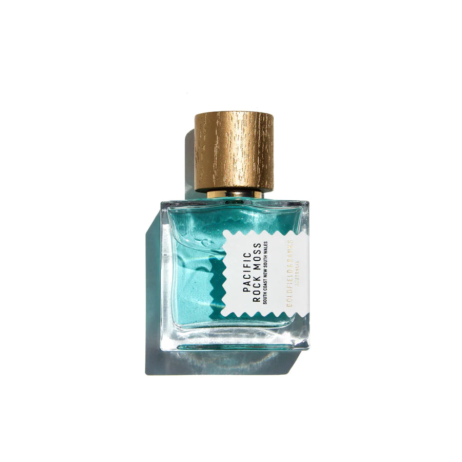 A clear glass bottle with a gold cap, labeled "Pacific Rock Moss" by Goldfield & Banks, houses a light blue liquid reminiscent of beach life. This fresh perfume exudes summer fragrance vibes. The label includes smaller text that is partially obscured by the bottle's angle and lighting.