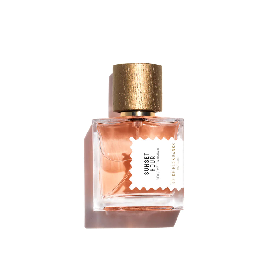 A square clear glass perfume bottle with a gold cap contains pink liquid. The label reads "Sunset Hour, Goldfield & Banks," evoking the serene beauty of an Australian sunset.