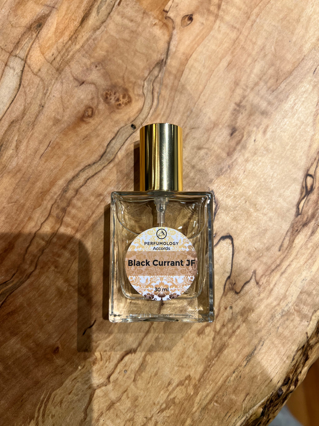 A 30 ml bottle of Perfumology Accords Black Currant JF perfume with a golden cap, exuding a fruity aroma, placed on a wooden surface.