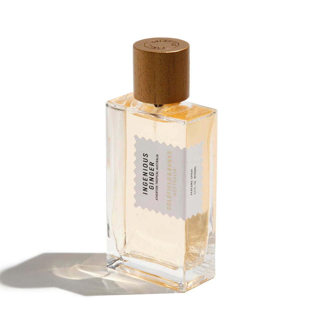 A clear glass perfume bottle with a wooden cap labeled "Ingenious Ginger" by Goldfield & Banks sits elegantly on a white background, its shadow lending depth. The tropical aroma promises an invigorating experience.